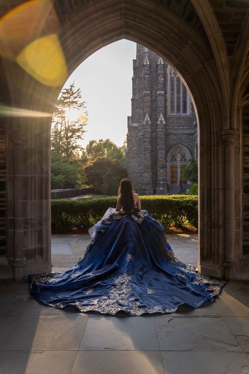 Quinceañera con vestido azul real de espaldas bajo un arco de piedra, con una capilla gótica al fondo