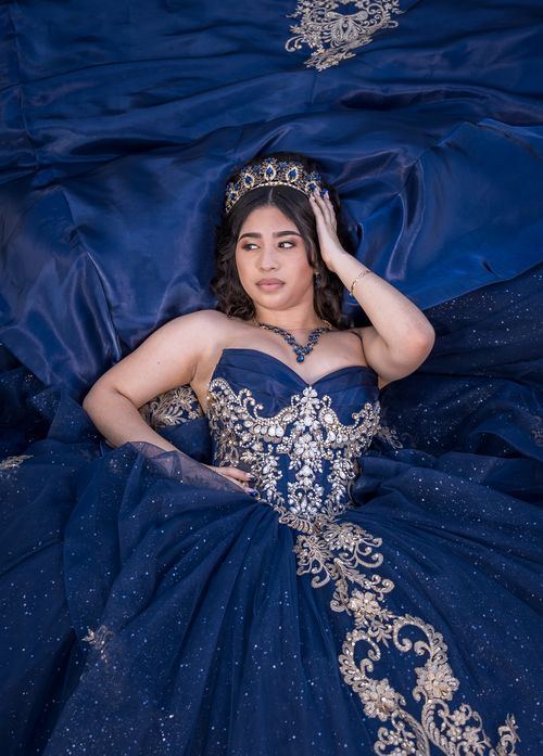 Quinceañera con corona recostada sobre la falda de su vestido azul bordado en plata